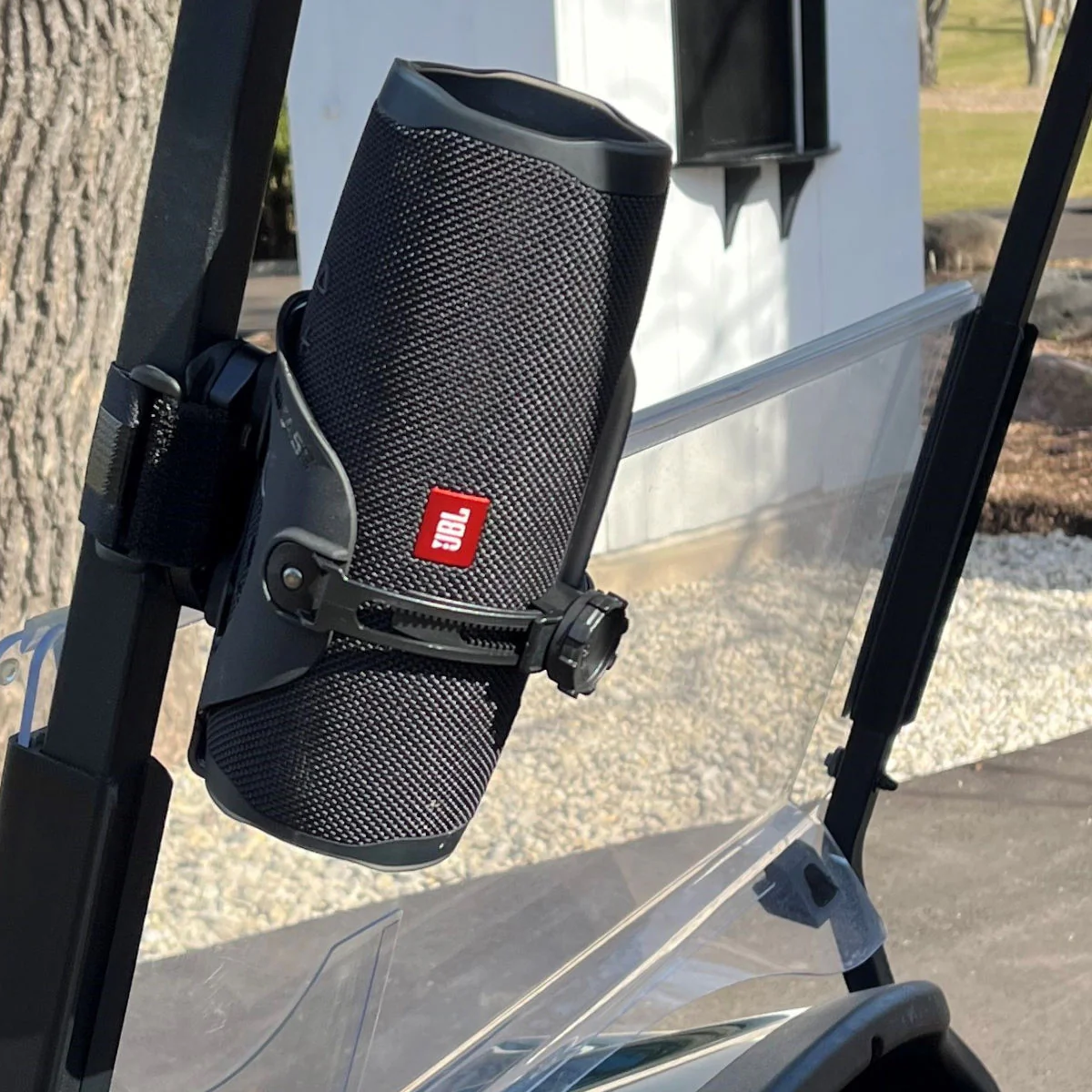 A black JBL portable speaker is mounted on the metal frame of a clear glass partition in an outdoor setting, showcasing universal compatibility similar to the Bikase Adjustable Drink Holder - BIKASE Anywhere.