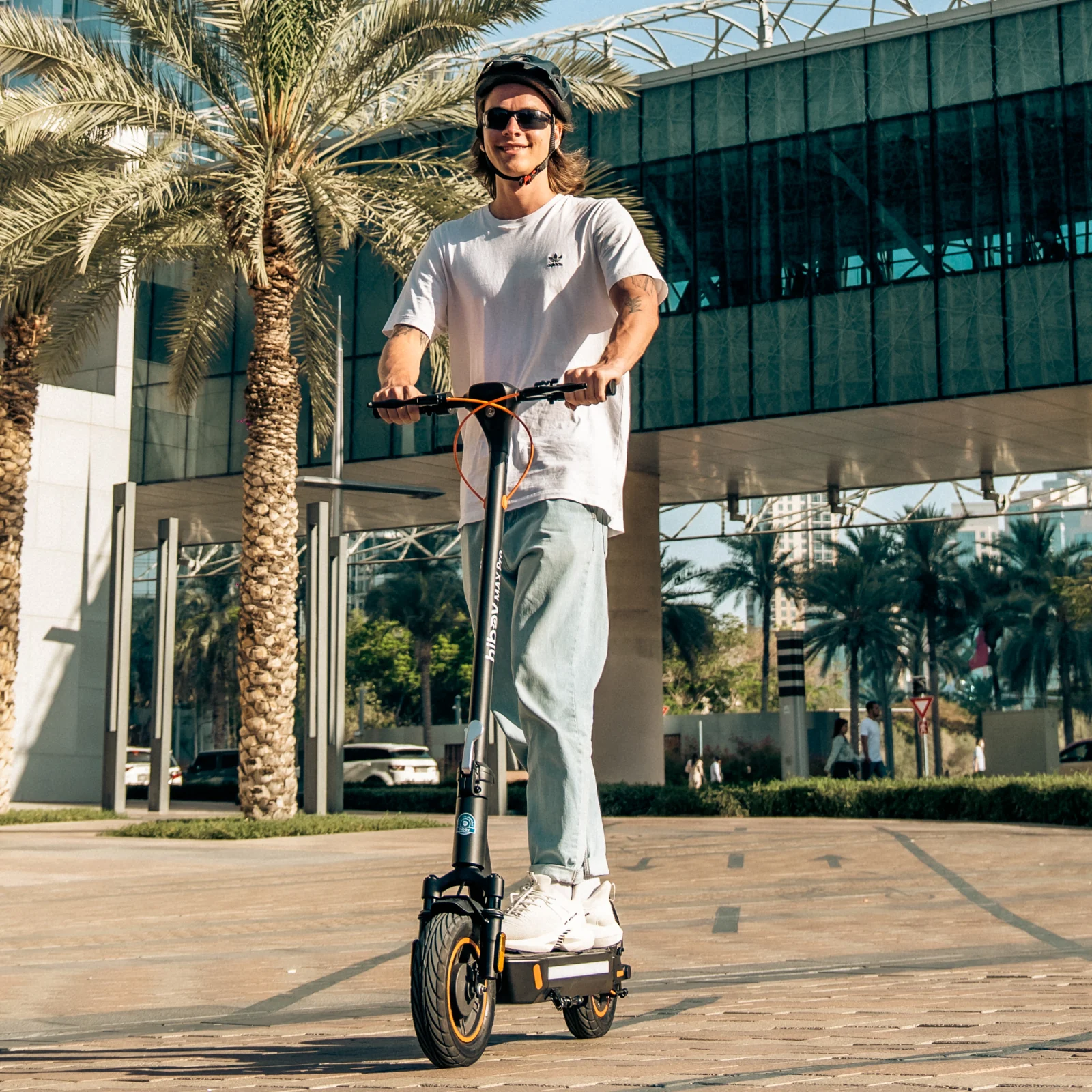 A person wearing a helmet, sunglasses, white t-shirt, and jeans rides the Hiboy MAX Pro Electric Scooter for Heavy Adult on a paved path lined with palm trees and modern buildings.