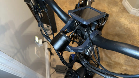 Close-up of a bicycle handlebar with a display screen, various cables, and a Tampa Bay eBikes Custom Headlight Mount Bracket securing the light.