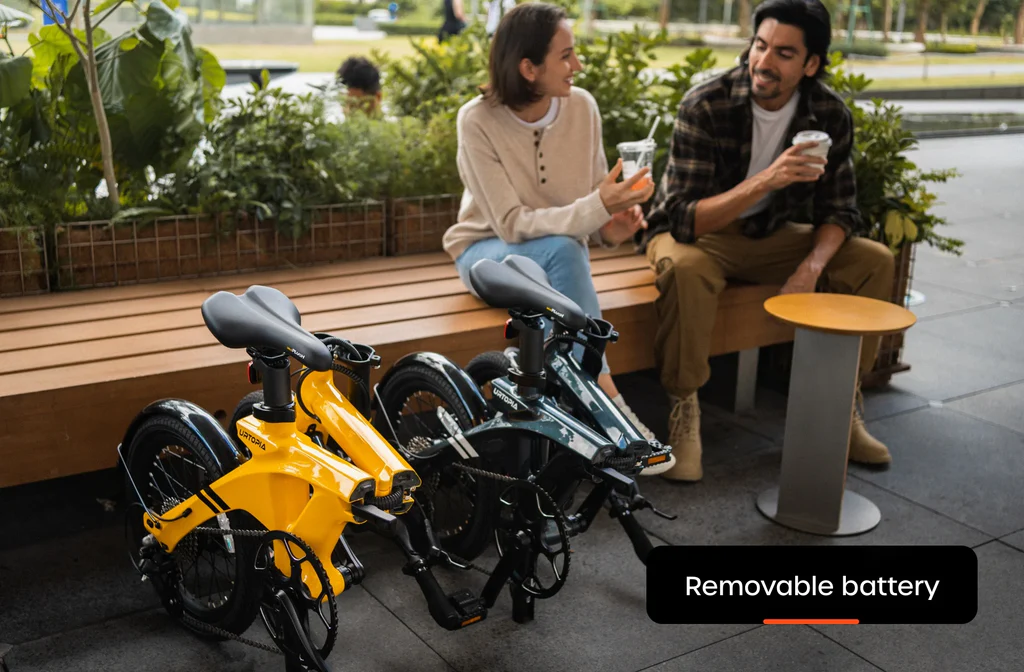 Two people sit outdoors talking on a bench, holding drinks, with two folded Urtopia Carbon Fold 1 eBikes by Urtopia parked in front; a label highlights the "Removable battery.