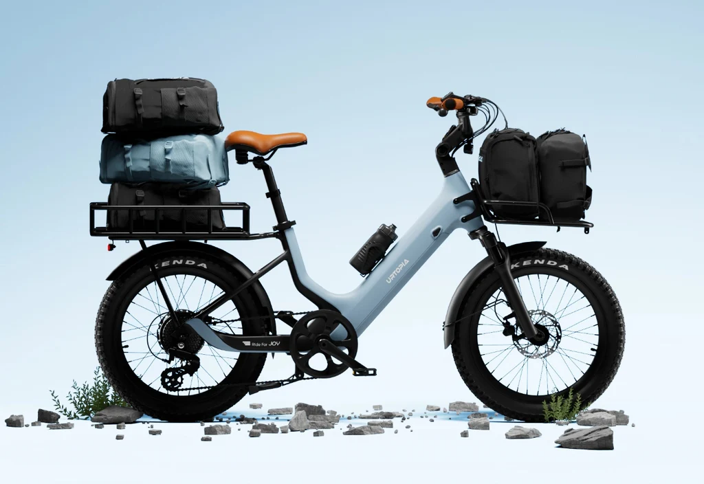 The Urtopia - Joy Carbon eBike by Urtopia, featuring a light blue frame, 750W motor, brown handlebars and seat, plus black panniers and bags on front and rear racks, stands on rocky ground with a pale blue background.