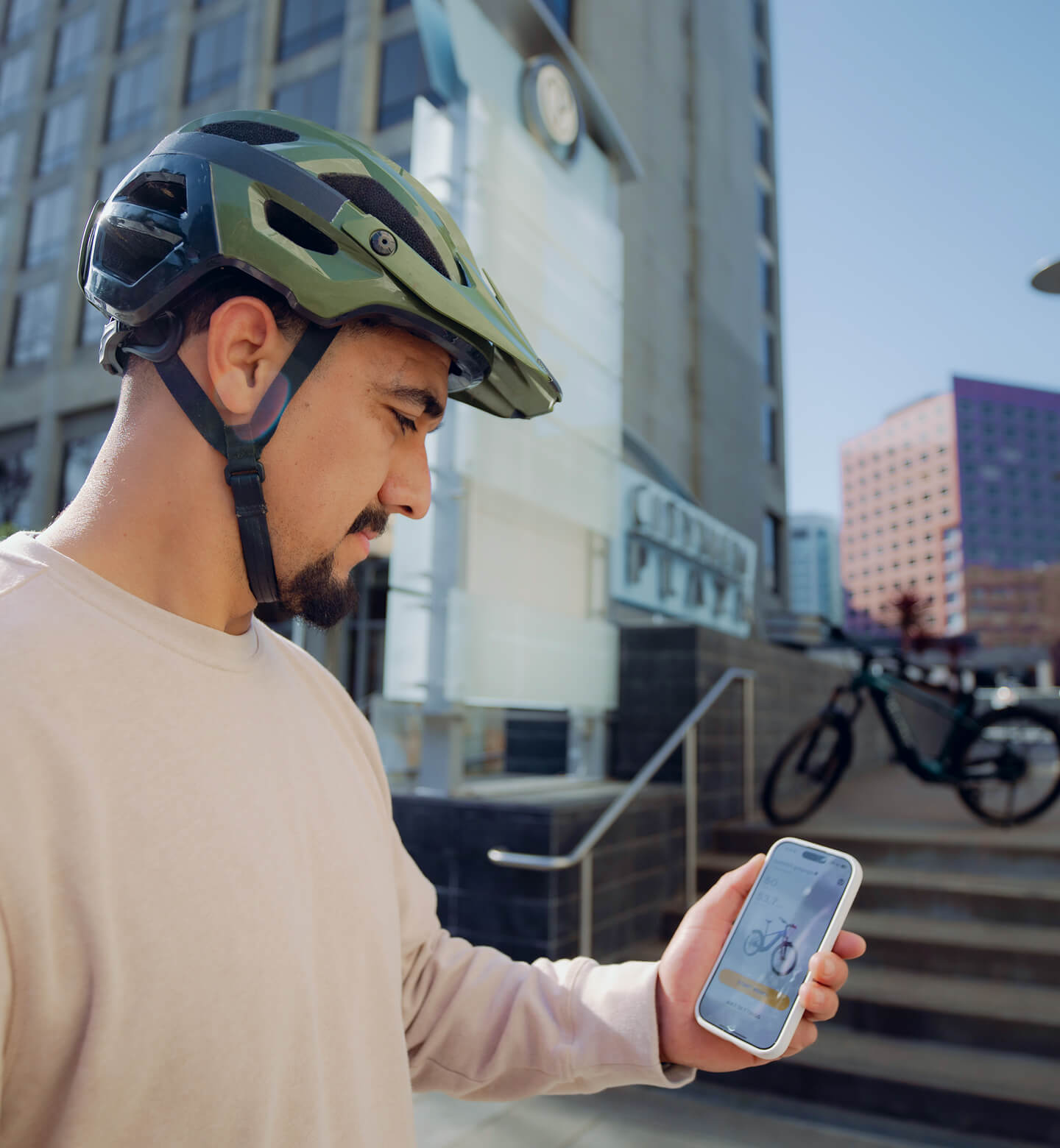 A man in a bike helmet stands outdoors, looking at his smartphone displaying the Velotric Summit 2 electric bike by Velotric, with buildings and a parked bicycle in the background.