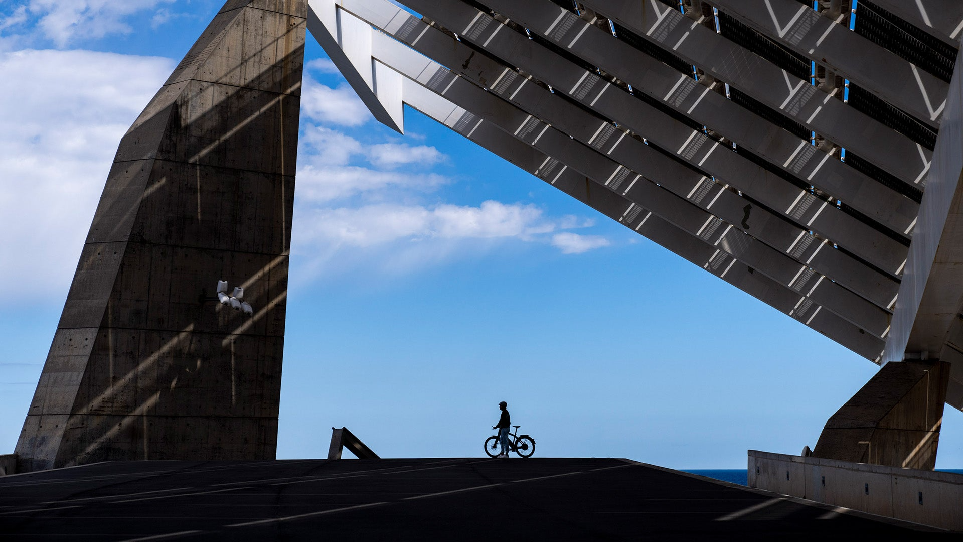 Silhouette of a cyclist riding a Stromer - ST7 Alinghi Red Bull Racing Edition eBike under a large, modern bridge with geometric structures and a clear blue sky in the background.