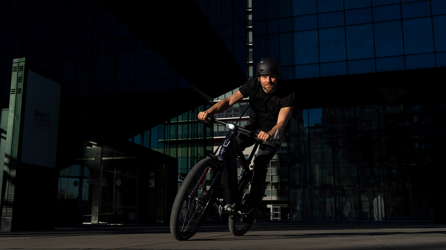 A man wearing a helmet rides a Stromer - ST7 Alinghi Red Bull Racing Edition eBike in front of a glass building with reflections, under a clear sky at sunset.