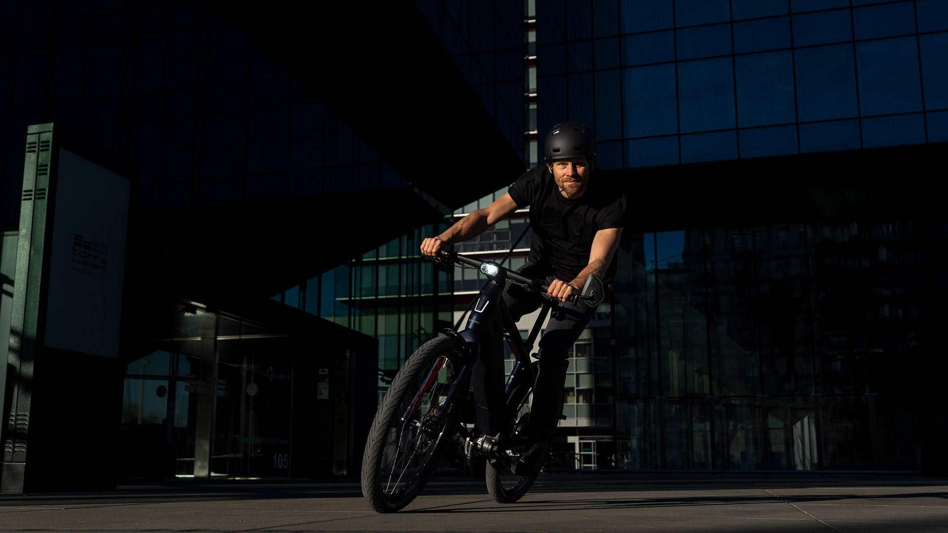A man wearing a helmet rides a Stromer - ST7 Alinghi Red Bull Racing Edition eBike in front of a glass building with reflections, under a clear sky at sunset.