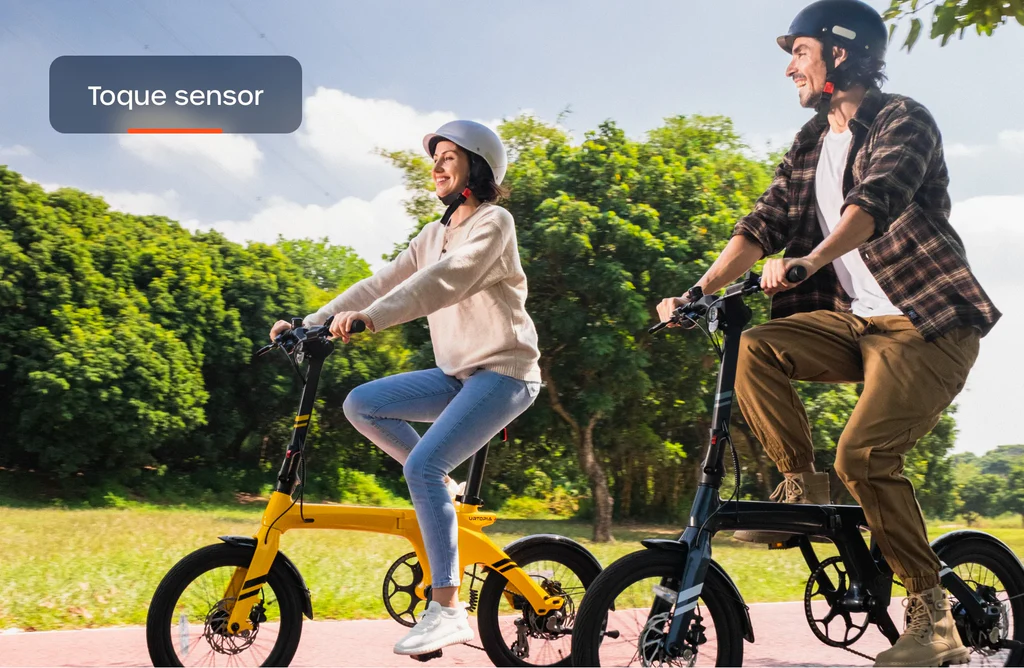 Two people wearing helmets ride Urtopia Carbon Fold 1 eBikes with carbon fiber frames along a tree-lined park path, enjoying the blue sky and scattered clouds.