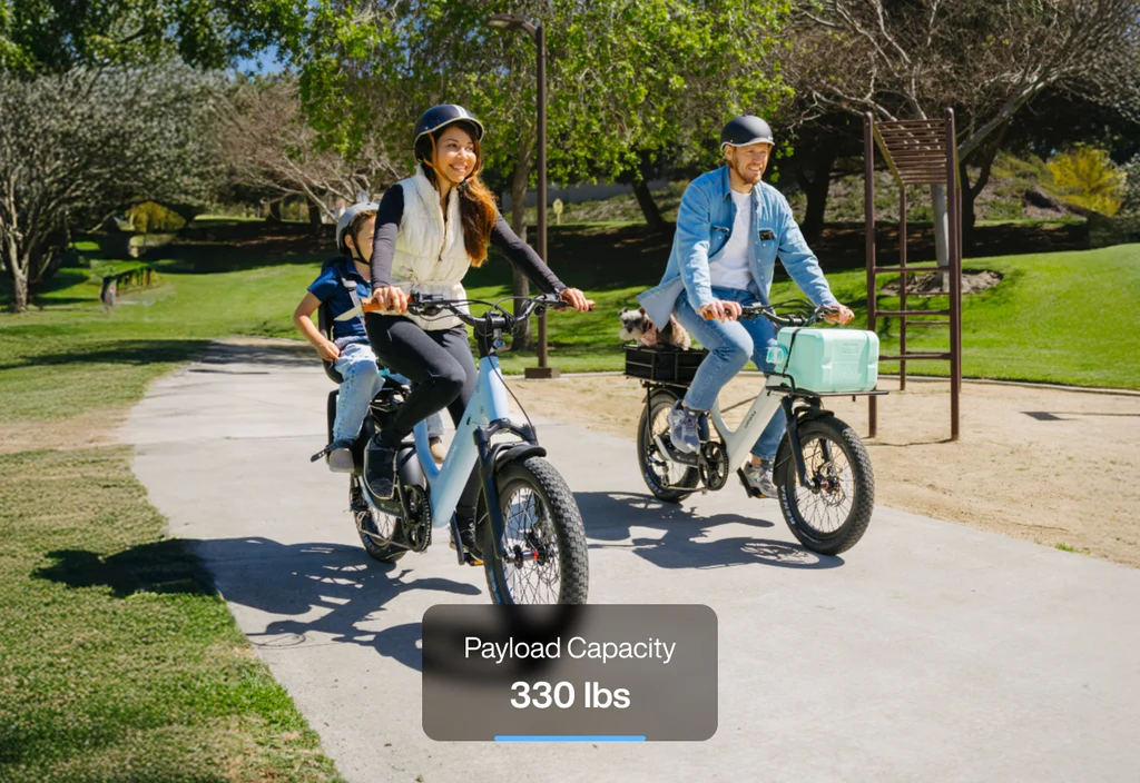 Two people ride Urtopia - Joy Carbon eBikes with strong carbon frames on a park path; the woman carries a child passenger. Text overlay reads "Payload Capacity 330 lbs.