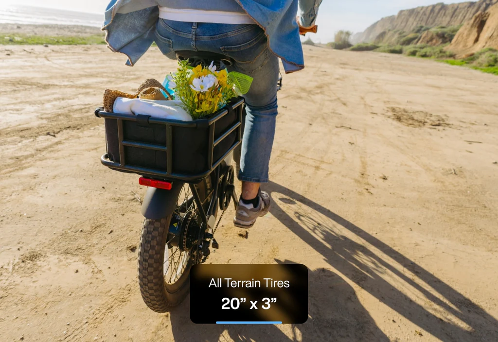 A person rides the Urtopia - Joy Carbon eBike by cliffs on a sandy path, carrying flowers and a bag in the rear basket. A label shows "All Terrain Tires 20” x 3”". Brand: Urtopia.
