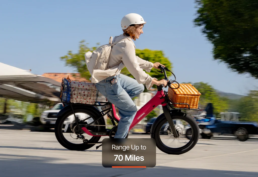 A rider in a helmet cruises on the Urtopia - Carbon Joy Ultra by Urtopia, a pink electric bike with front and rear baskets. Text overlay: "Range Up to 70 Miles.
