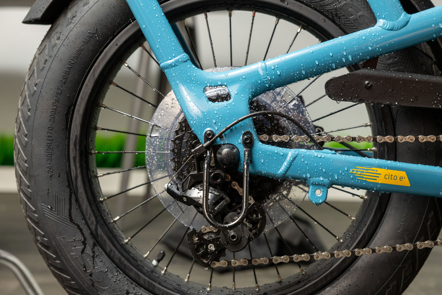 Close-up of a wet blue Momentum - Cito E+ bicycle rear wheel with a detailed view of the chain, gears, and tire tread, highlighting water droplets.