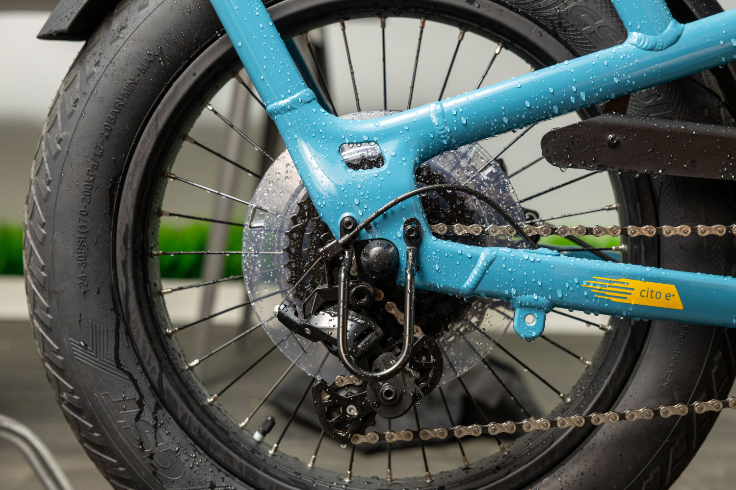 Close-up of a wet blue Momentum - Cito E+ bicycle rear wheel with a detailed view of the chain, gears, and tire tread, highlighting water droplets.