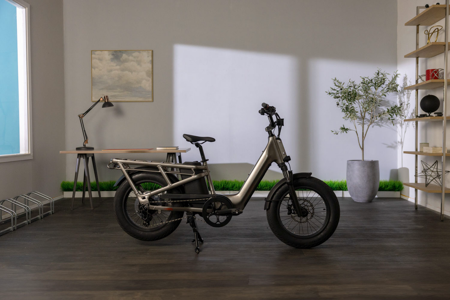 Momentum - Cito E+ electric bicycle, parked in a modern home office with a desk, lamp, shelving units, and plants, illuminated by natural light from a window.