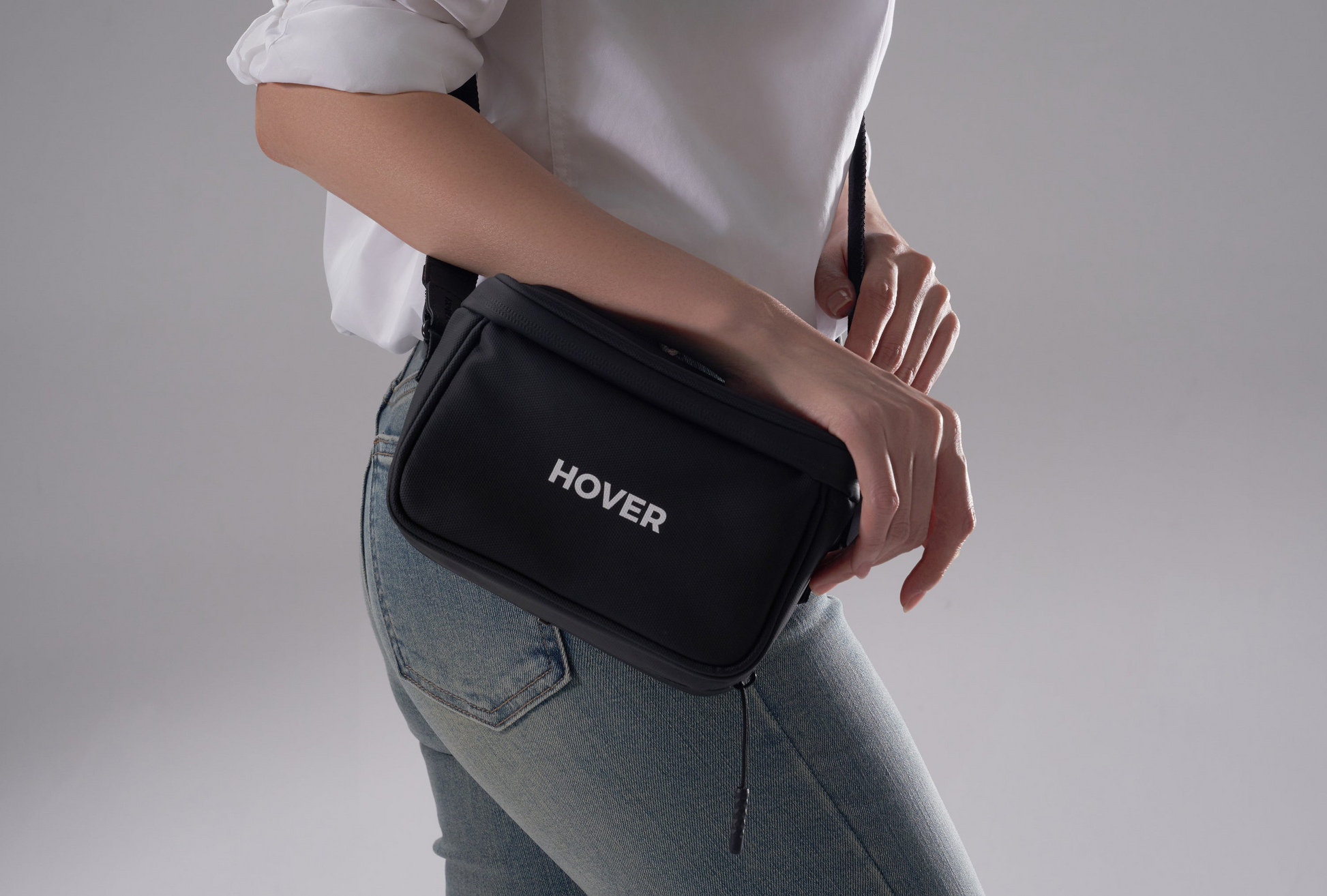 A person in jeans and a white shirt holds a HOVERAir Carry Bag, black with "HOVER" branding, designed to complement the HoverAir X1 Pro lifestyle against a plain background.