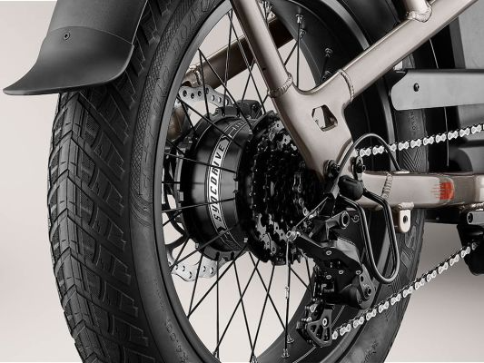Close-up of a Momentum - Cito E+ rear wheel assembly, showing details of the tire, chain, brake system, and suspension components against a neutral background.