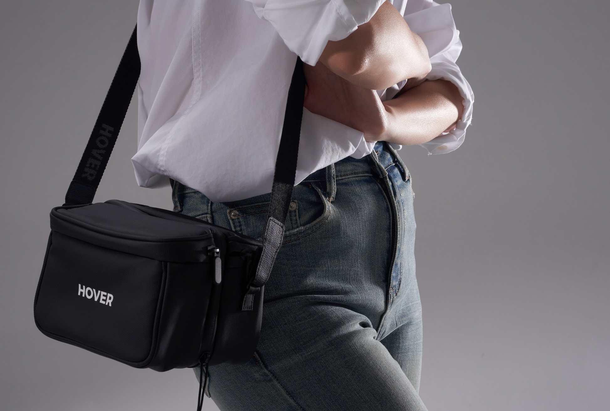 A person in a white shirt and jeans confidently stands against a gray background, wearing a black HOVERAir Carry Bag with the "HOVER" label across their body.