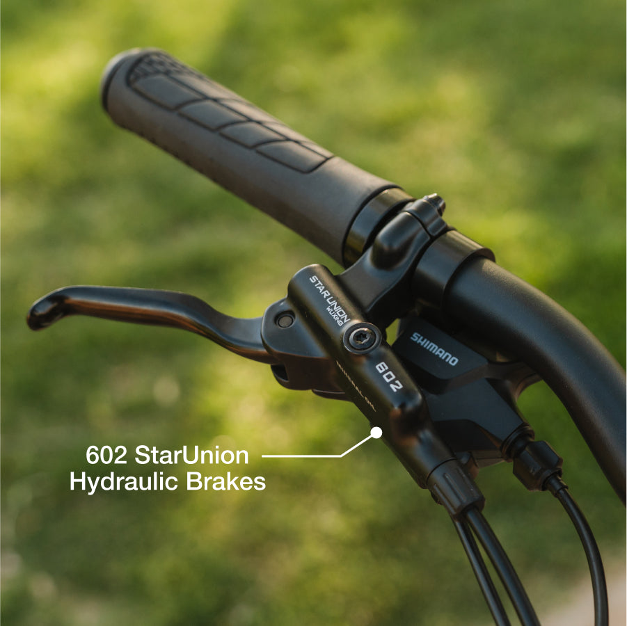 Close-up of the Lectric XP4 folding eBike handlebar with Shimano components and "602 StarUnion Hydraulic Brakes" label, set against a blurred green background. Powered by a 750W rear hub motor from Lectric for enhanced performance.