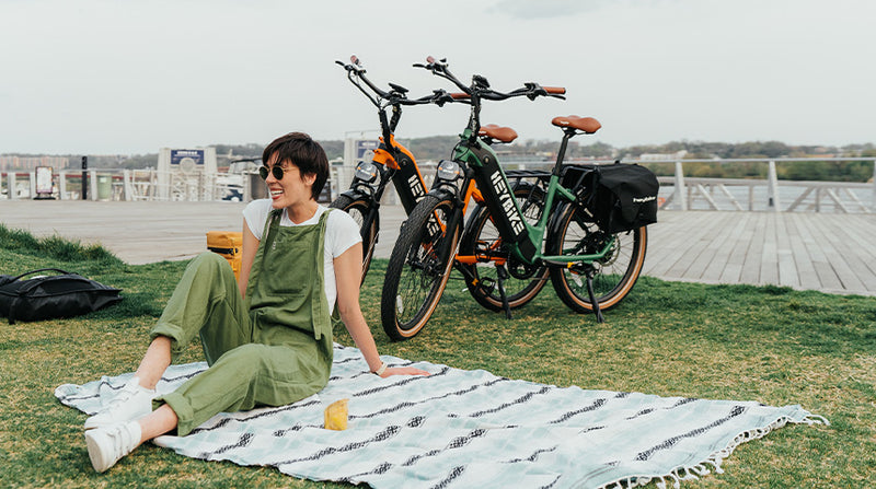 Woman in green overalls sitting on a picnic blanket outdoors, smiling, with two HeyBike Cityrun ebikes parked behind her on a wooden pier.