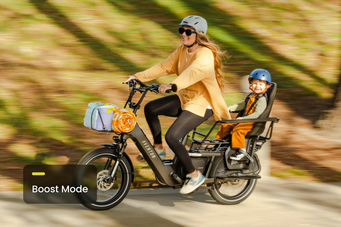 An adult and child, both with helmets, ride the Aventon Abound LR cargo e-bike from Aventon on a sunny day. A "Boost Mode" label appears on the image.