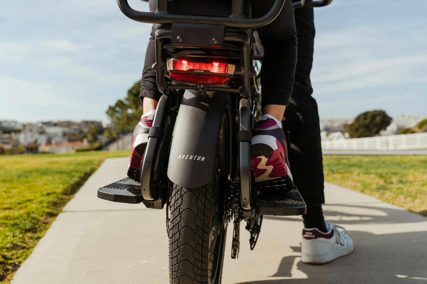 A person in sneakers sits on the rear of an Aventon Abound LR cargo e-bike by Aventon, with its rear wheel and brake light visible while parked on a paved path in a park.