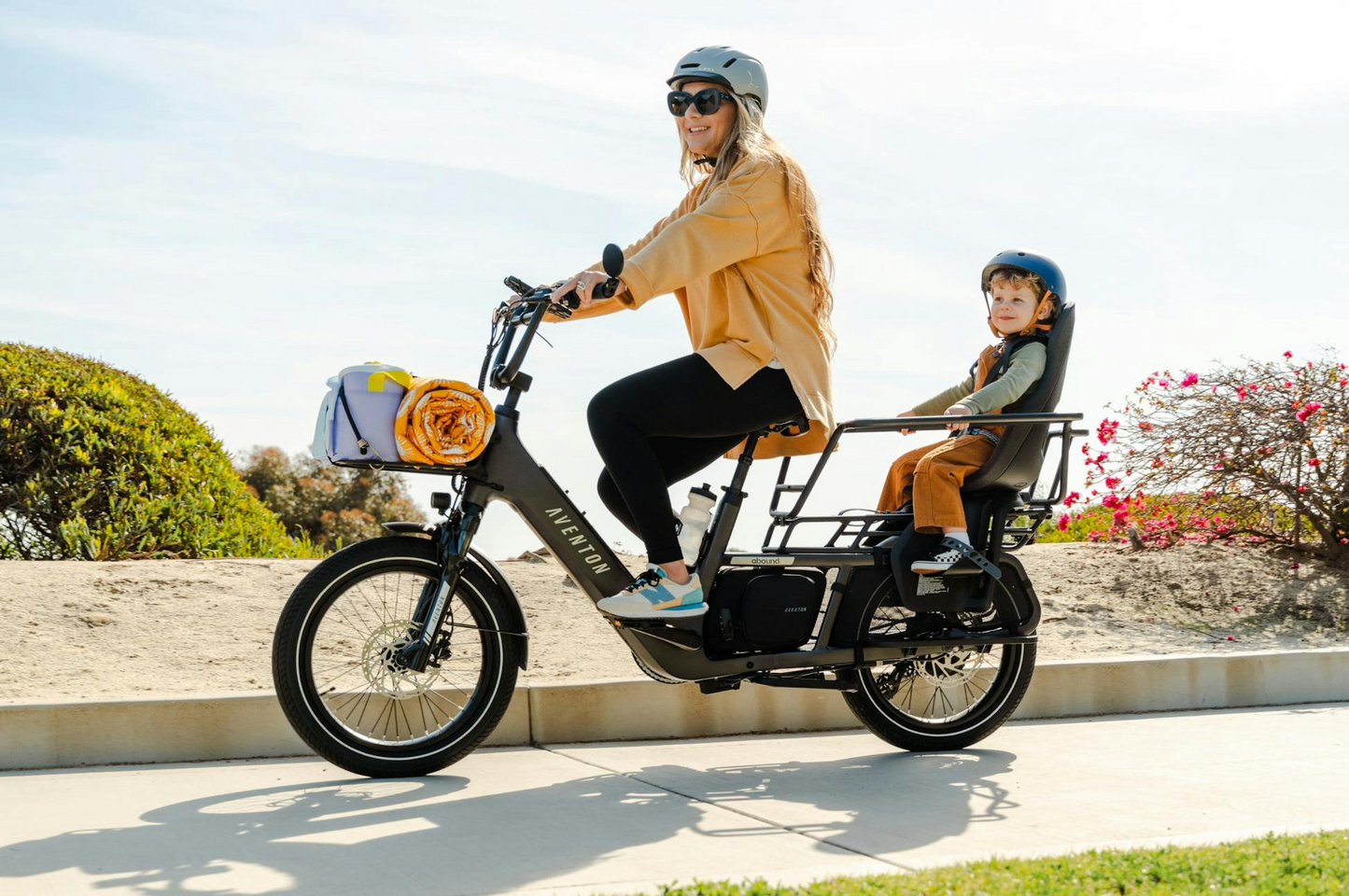 A helmeted woman rides an Aventon Abound LR cargo e-bike with a smiling child in the rear seat; both enjoy a sunny day outdoors.
