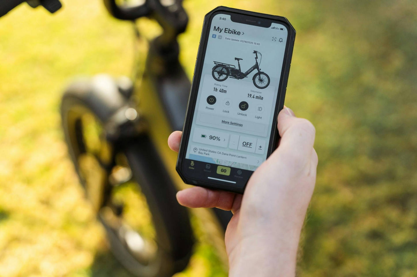 A person holds a smartphone displaying an e-bike app with battery status and ride info, while the Aventon Abound LR electric cargo bike by Aventon appears blurred in the background.
