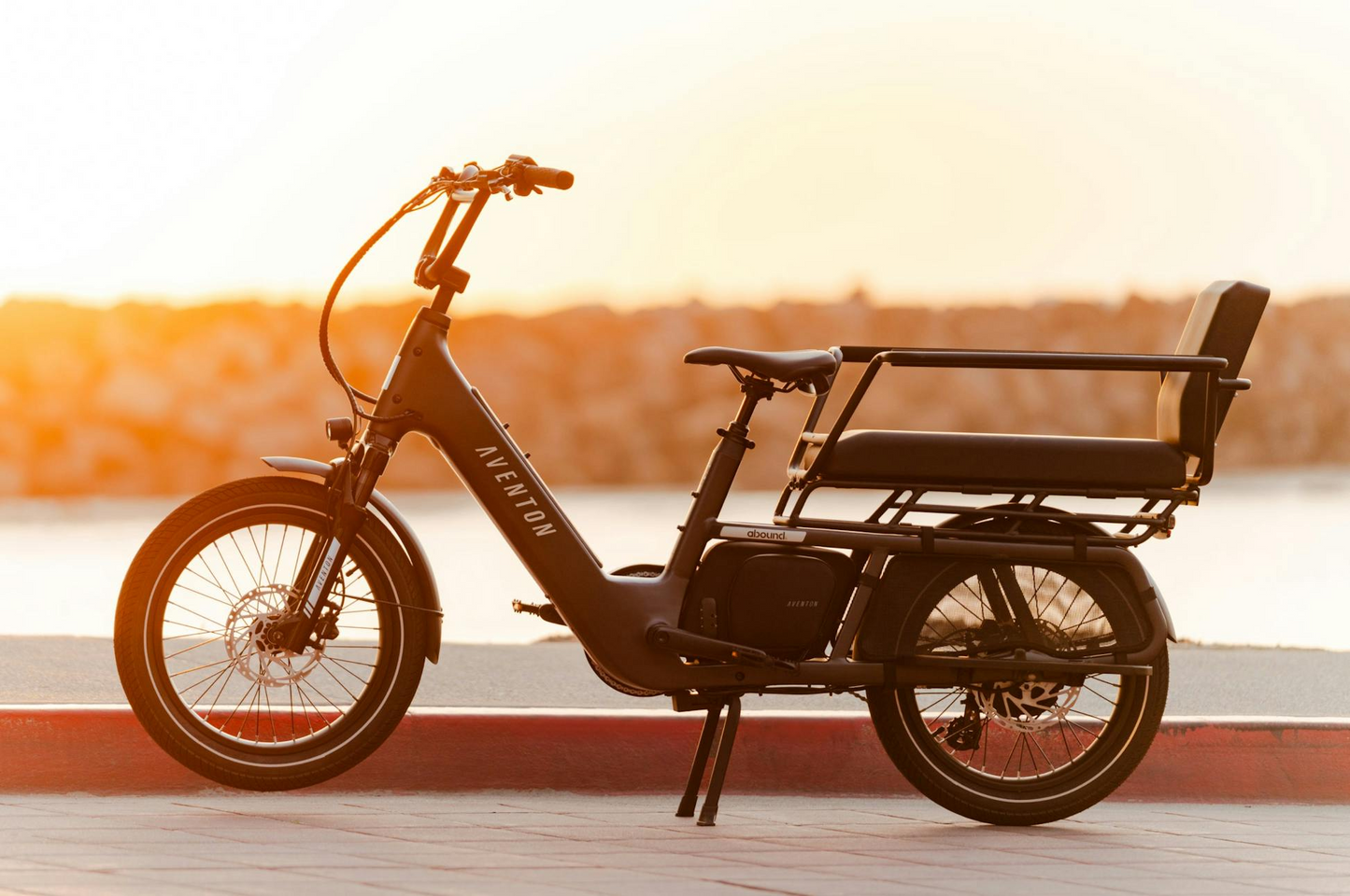 A black Aventon Abound LR electric cargo bike from Aventon with a rear bench seat is parked on a sidewalk near the water at sunset.