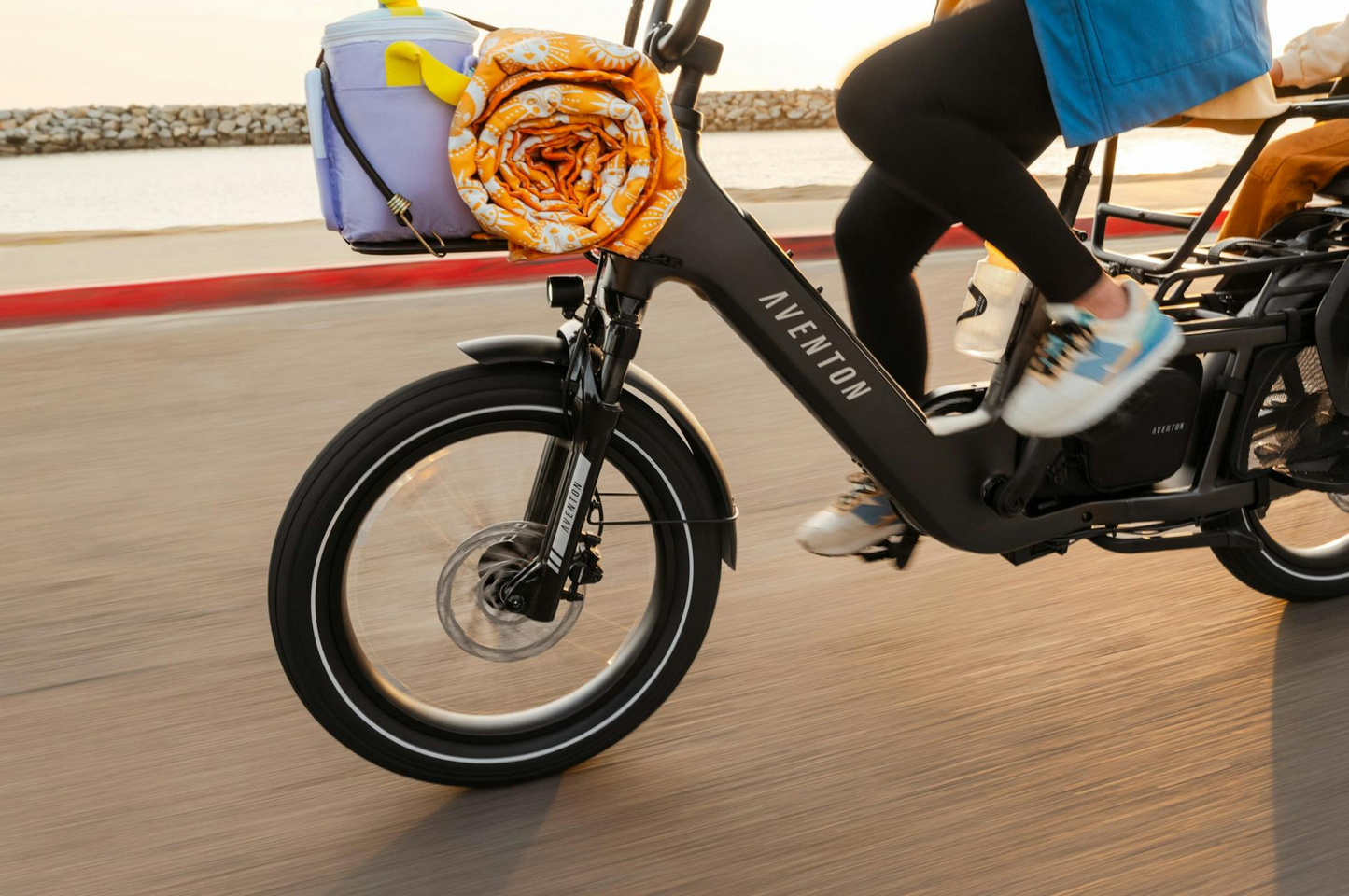Someone rides the Aventon - Abound LR electric cargo bike by the water, carrying a towel and cooler on its front rack.