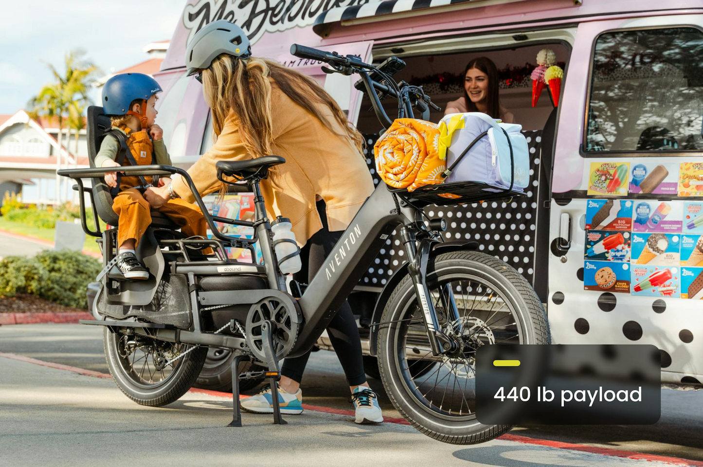A woman assists a child seated on an Aventon Abound LR by Aventon cargo e-bike, parked near an ice cream truck, with a 440 lb payload capacity label visible.
