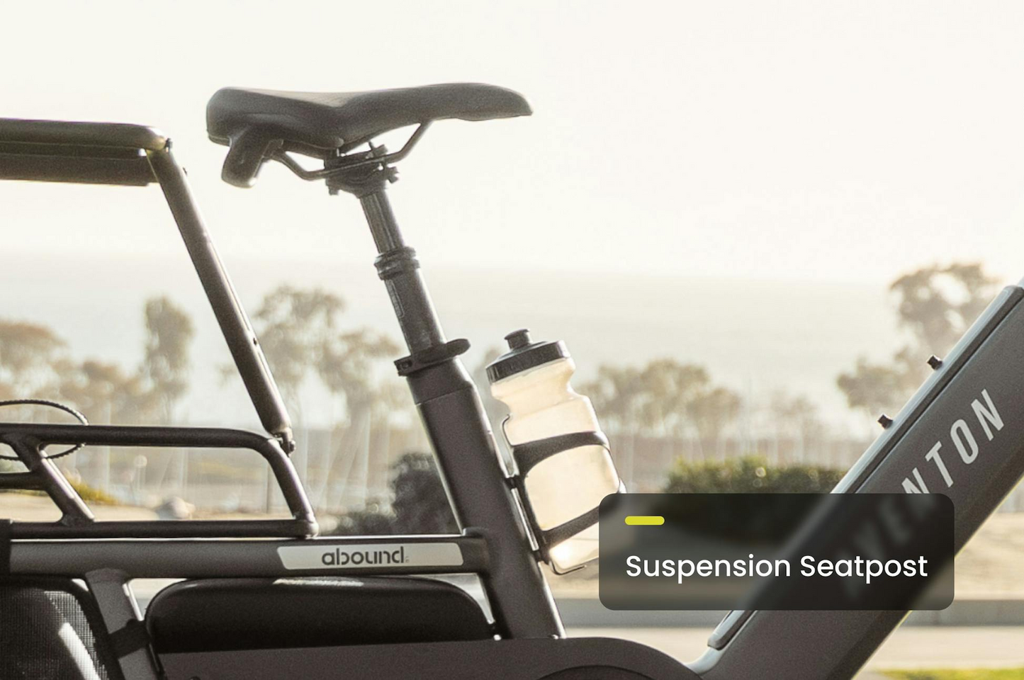 A close-up of the Aventon - Abound LR cargo e-bike by Aventon features its suspension seatpost, water bottle, and rear rack with softly blurred trees and water in the background.