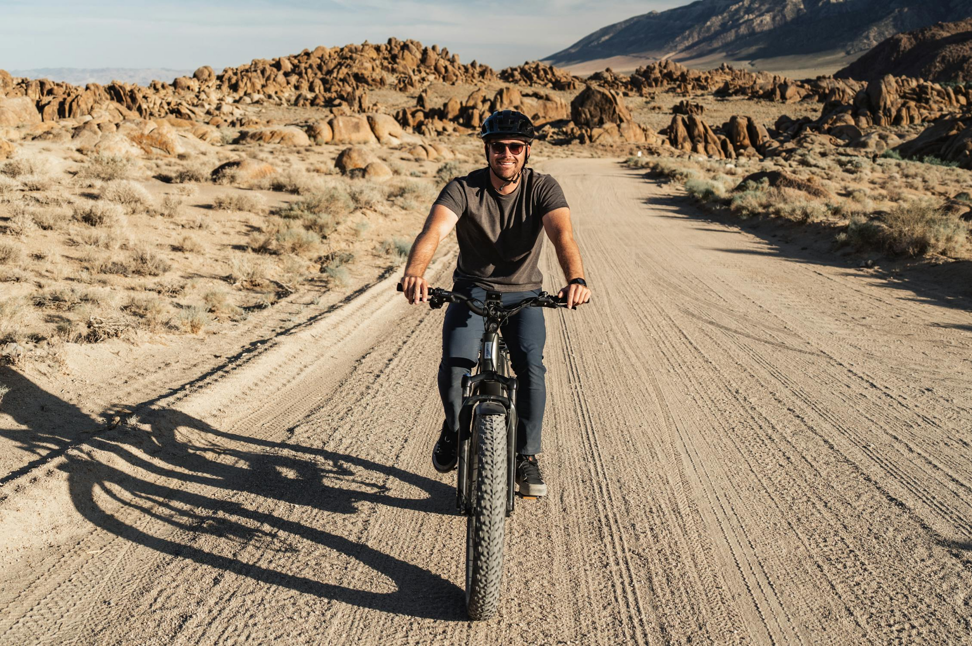 A person wearing a helmet rides a Tampa Bay eBikes Aventon - Aventure M fat tire eBike on a dirt road through a rocky desert landscape under a clear sky.