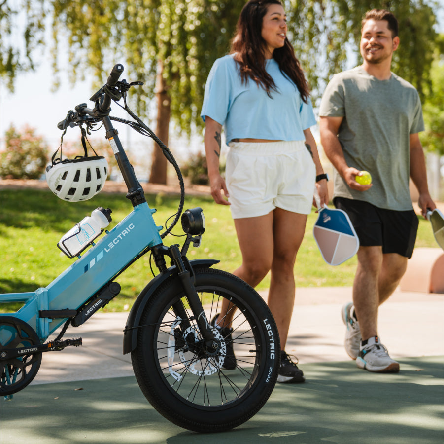 Two people walk outside on a sunny day holding pickleball paddles and a ball, while a blue Lectric XP4 folding eBike by Lectric is parked in the foreground.