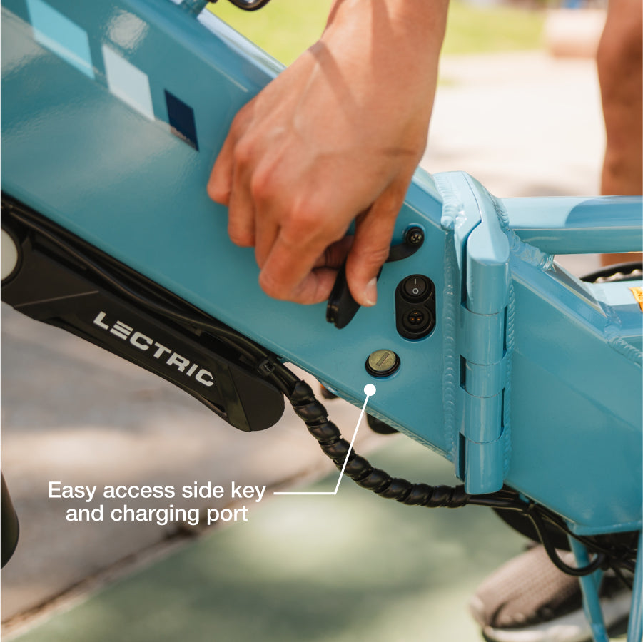 A hand connects a charger to the side charging port of a blue Lectric XP4 folding eBike; labels indicate the key and charging port.