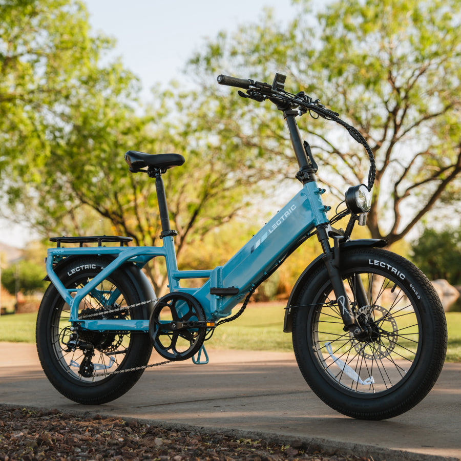 A blue Lectric XP4 folding eBike with a 750W rear hub motor is parked on a paved park path, surrounded by trees and grass.