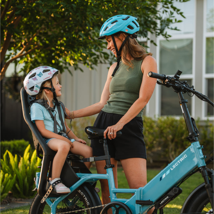 A woman and a child, both in helmets, smile at each other while seated on a blue Lectric XP4 folding eBike with a powerful 750W rear hub motor, by Lectric, in a residential neighborhood.