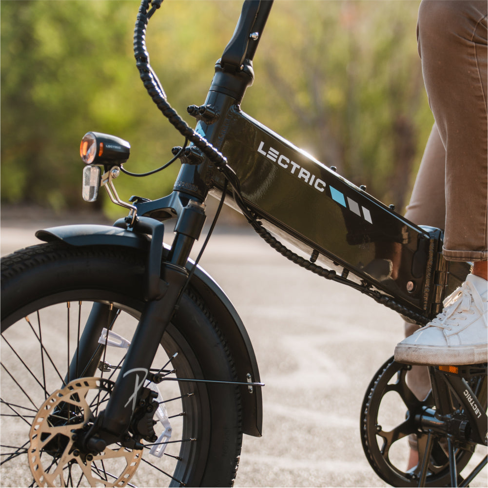 A person in brown pants and white shoes rides the Lectric XP4, a black folding eBike by Lectric with a 750W rear hub motor, along a paved path surrounded by greenery.