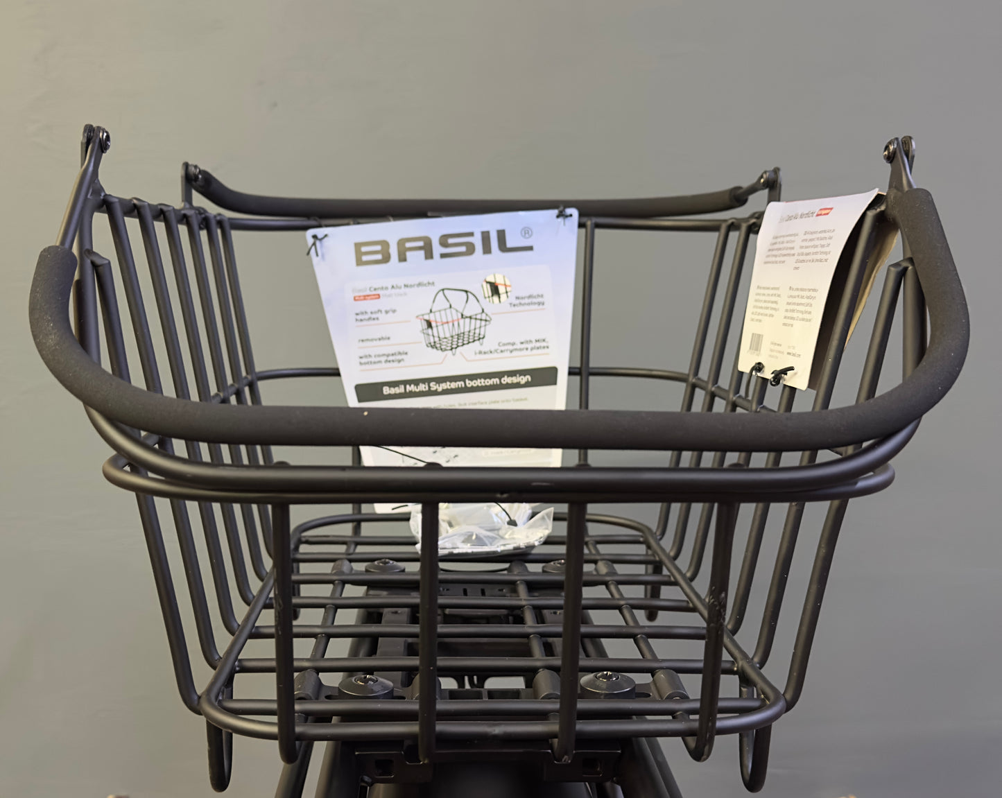 The Basil Cento MIK Rear Basket, a black metal bike basket with a "Basil" label and instruction sheet attached, is shown mounted on a bicycle against a plain gray background.