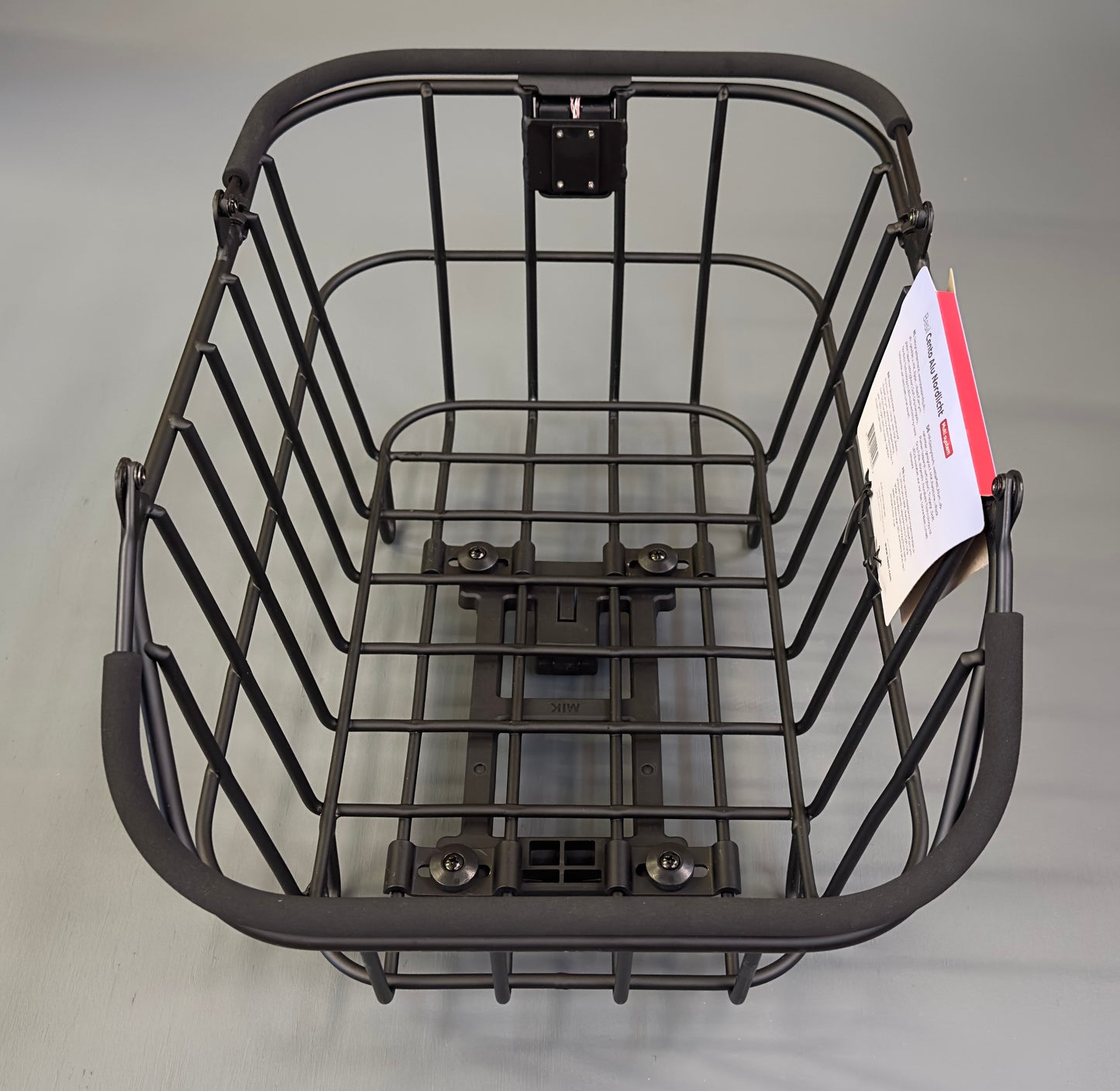 The Basil Cento MIK Rear Basket is a black metal wire e-bike basket with a mounting bracket at the base and a paper tag attached. Empty and placed on a plain surface, it is compatible with the MIK system for easy e-bike installation.