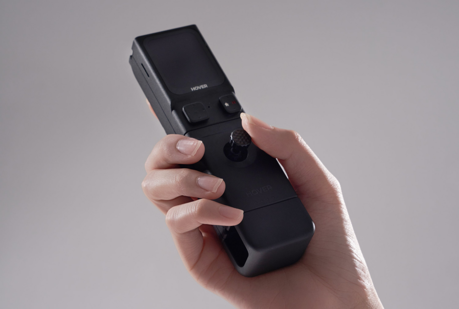 A hand holds the sleek black HOVERAir Beacon and JoyStick Bundle, featuring a small screen and joystick with cutting-edge HoverLink technology, set against a plain background.
