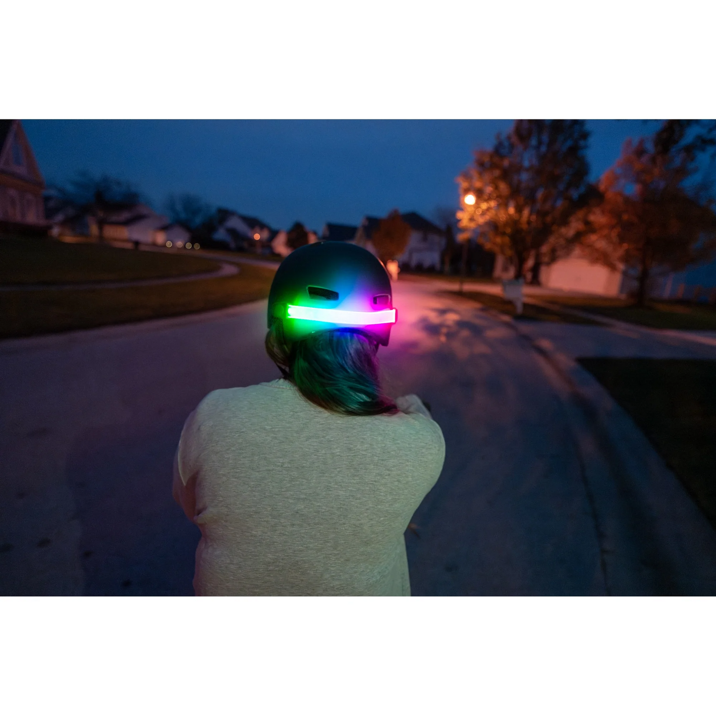 A person is wearing the Brightz Helmet Brightz, featuring colorful LED lights and a color select ring on the back, while standing on a residential street at dusk.