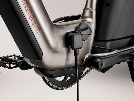 Close-up of a Momentum - Cito E+ mid-frame, highlighting the charging port connected to a charger, with a clear view of the chain and pedal.