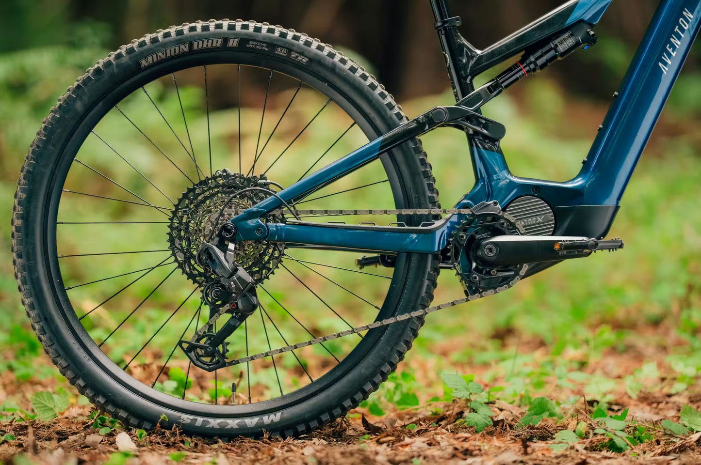 A close-up of the rear wheel, gears, and pedal of the blue Aventon Current ADV full-suspension eMTB by Aventon on a forest floor with green plants and fallen leaves.