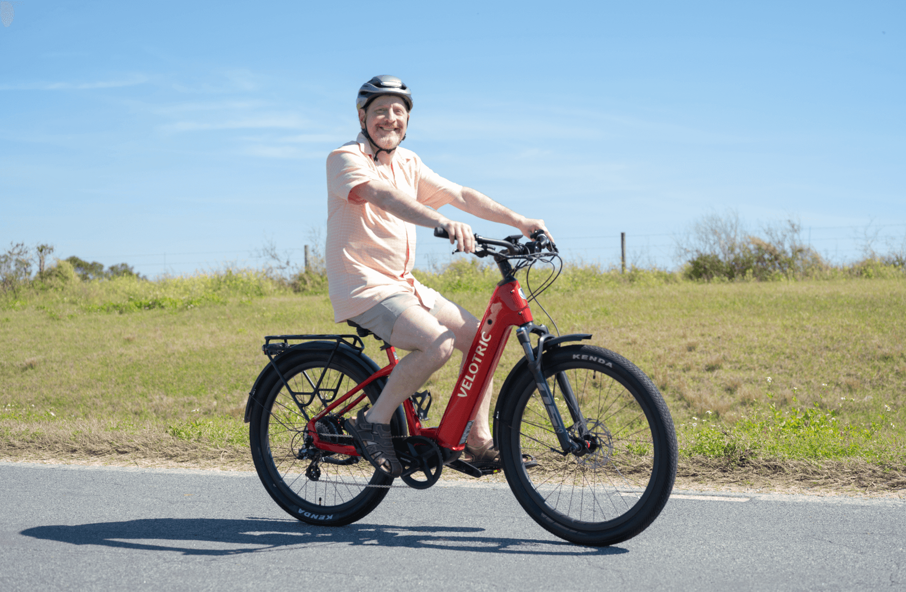 Man riding a red Velotric - Discover 1 on a paved road with a grassy field in the background. He is wearing a helmet and light-colored clothing, exemplifying the Tampa Bay eBikes style.