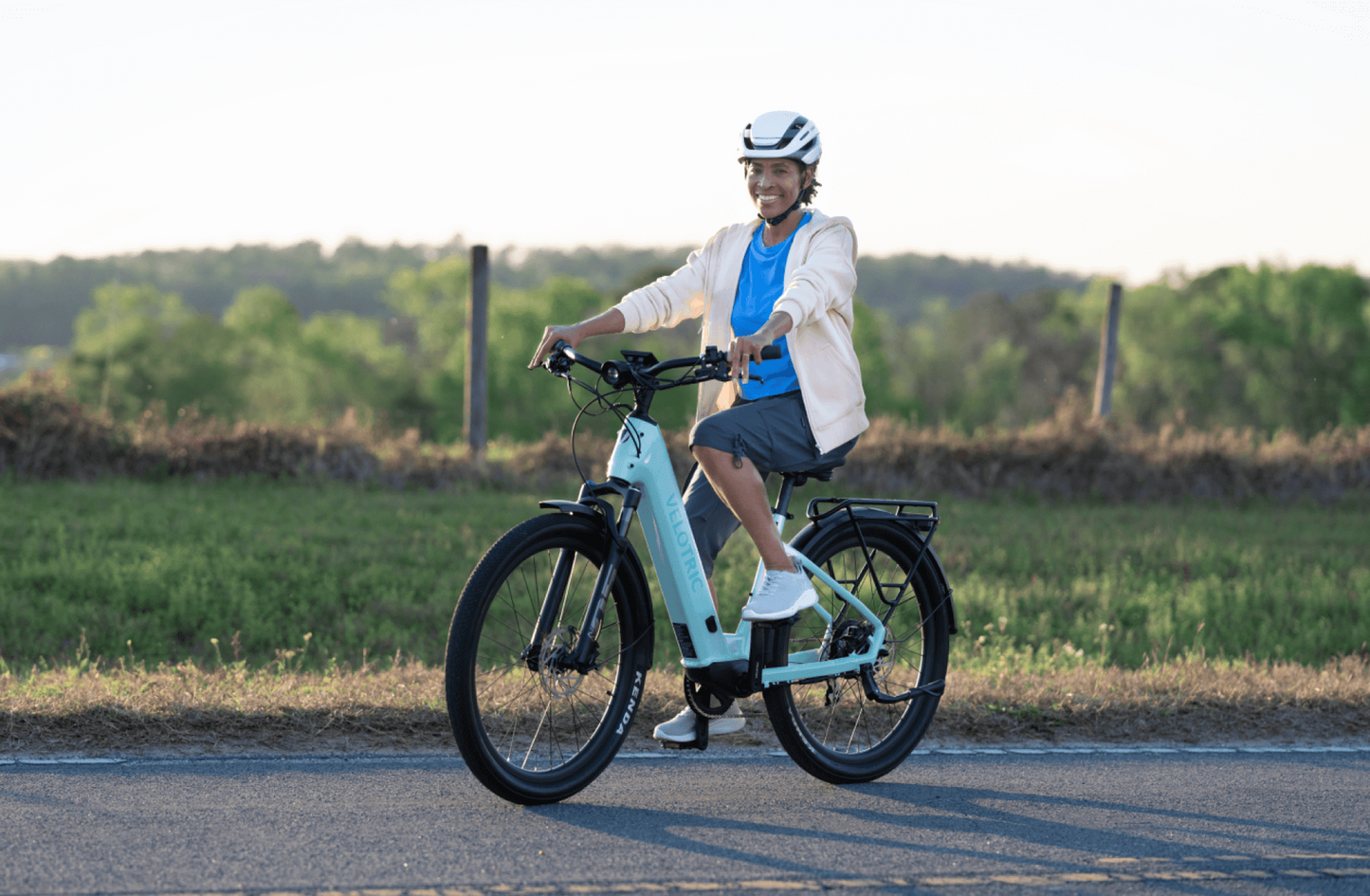 A person in a helmet and casual clothing is smiling while seated on a stationary Velotric - Discover 1 e-bike on a road with greenery in the background.