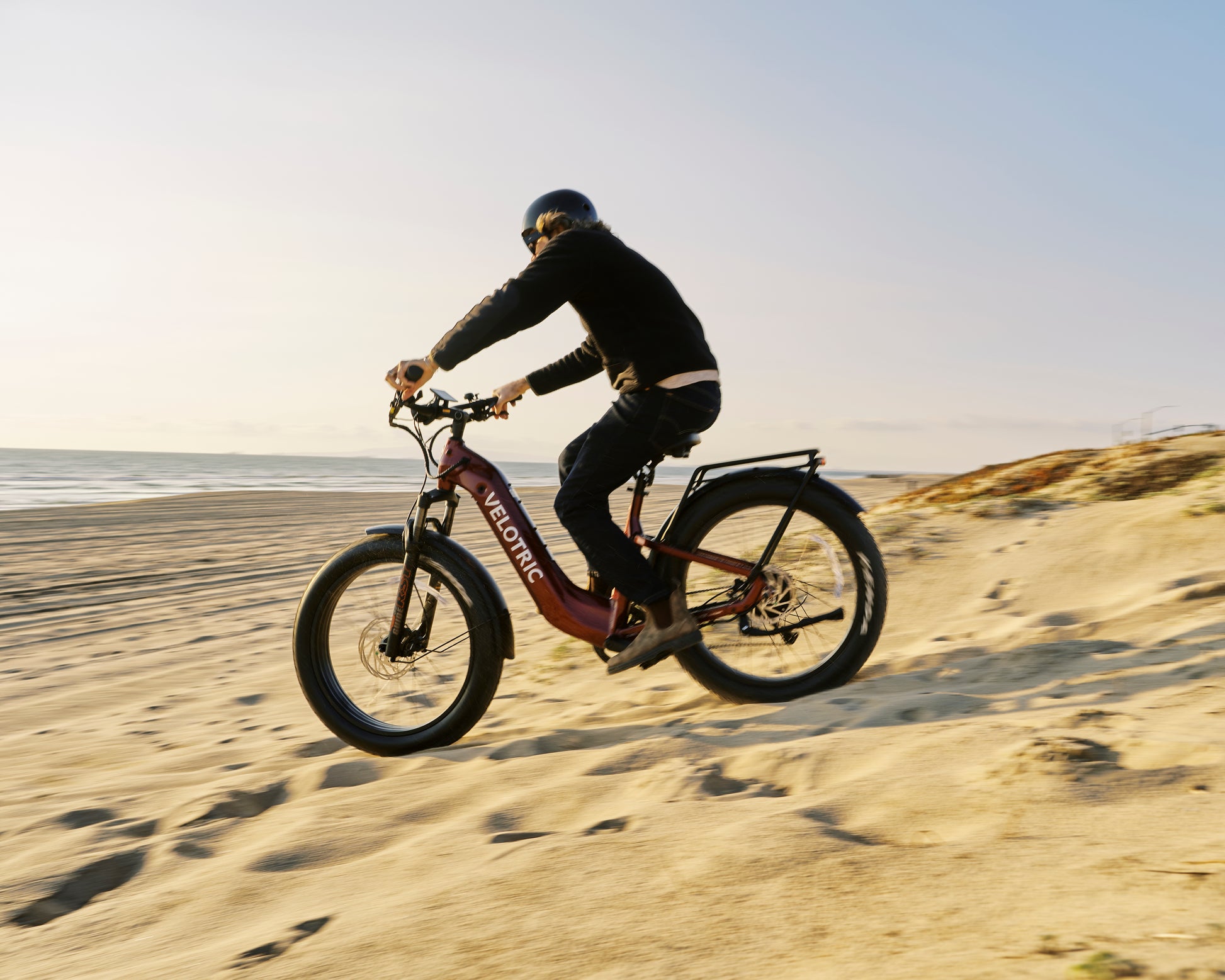Riding the Velotric - Nomad 2, a person effortlessly navigates the sandy beach; its all-terrain capability and fat tires glide smoothly against the ocean backdrop.