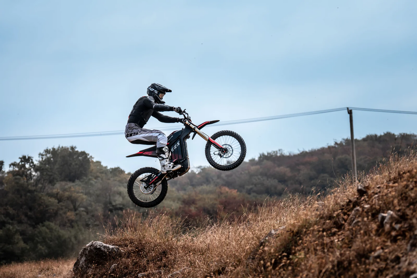 A rider in protective gear takes the Navee Storm X electric dirt bike by Navee uphill over grassy, rocky terrain with trees and clear skies, highlighting its impressive off-road performance.