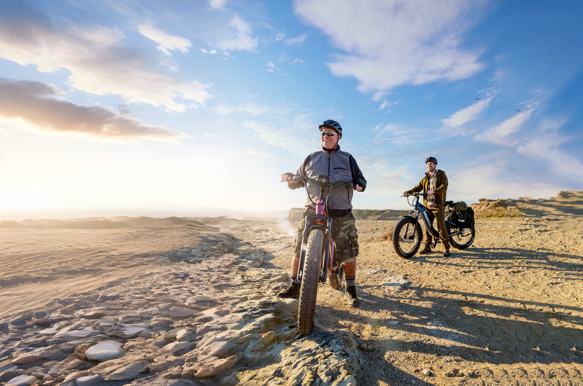 Two adventure seekers with Velotric - Nomad 2 bicycles stand on rocky terrain under a partly cloudy blue sky, wearing helmets and casual outdoor clothing.