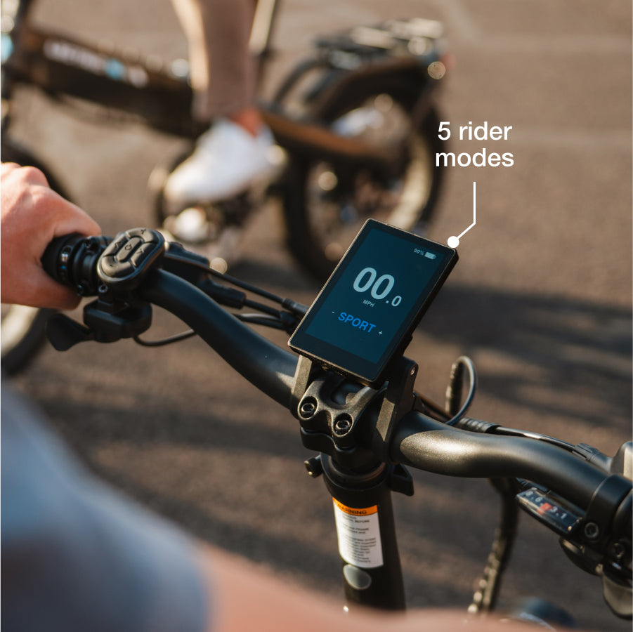Close-up of a person’s hands on Lectric XP4 handlebars, displaying "00.0 SPORT" and "5 rider modes." This folding eBike by Lectric features advanced Torque Sensor Technology, with another cyclist in the background.