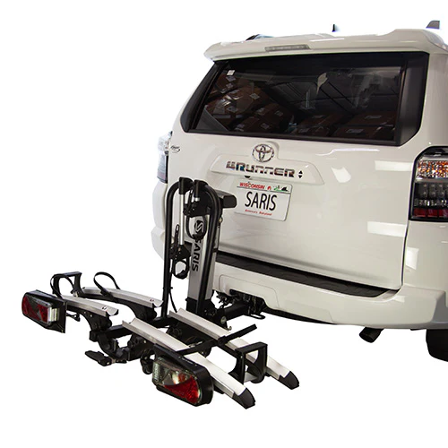 A white SUV with a rear-mounted Saris - Door County 2-Bike Motorized Hitch Rack from Hollywood Racks securely carries two eBikes. The vehicle, featuring a "SARIS" license plate, is parked indoors, highlighting its efficient eBike transport solution.