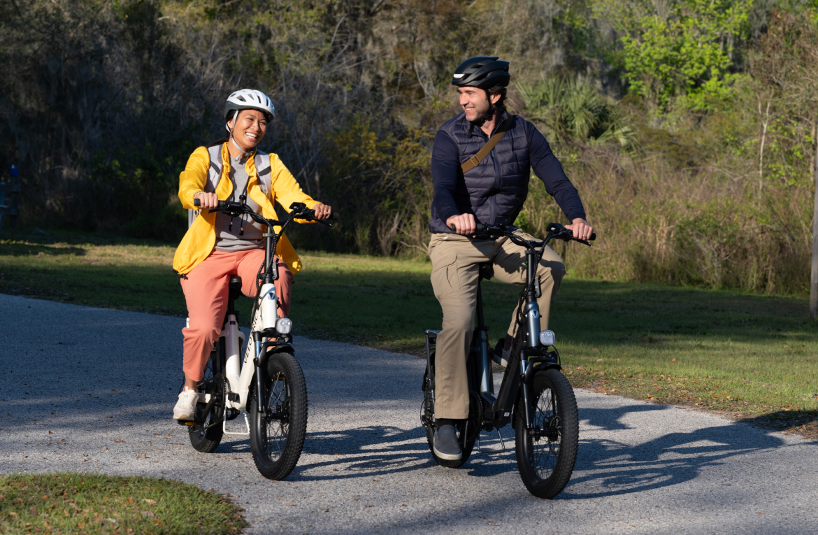 Two people riding Velotric - Fold 1 eBikes on a sunny park path, smiling and enjoying a leisurely ride together.