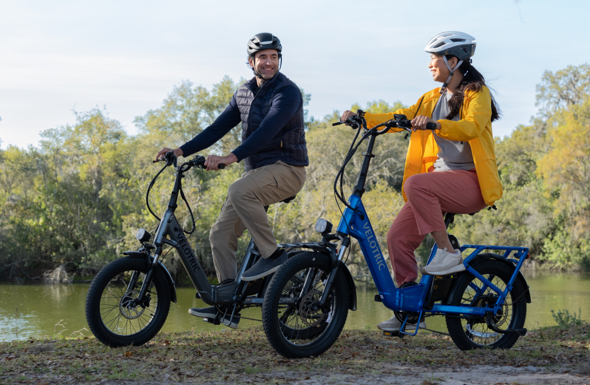 Two people riding Velotric Fold 1 bikes by a river in a sunny park, smiling at each other, wearing helmets.